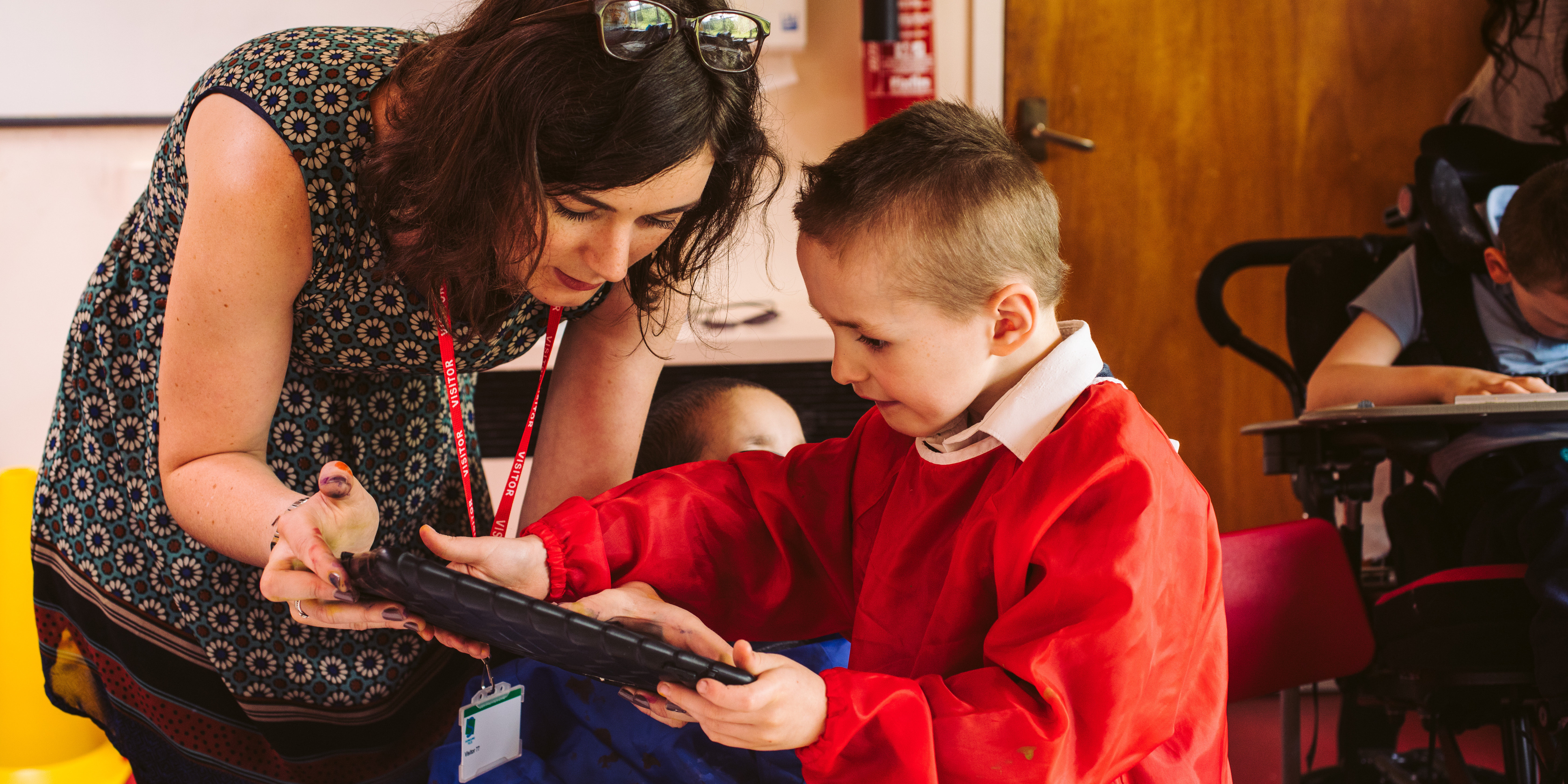A female artist in a classroom holds an ipad for a primary aged boy in a red art smock
