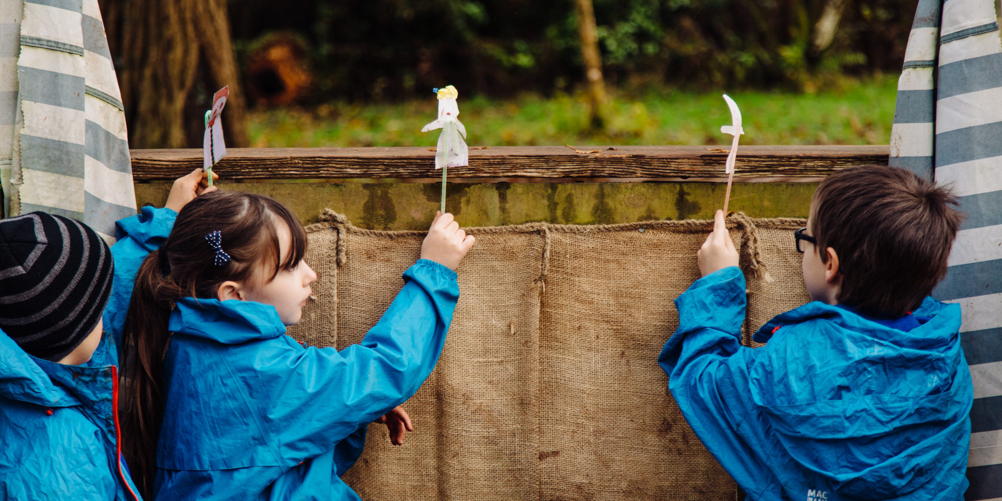 Three primary aged children in blue macs lift up three puppets on sticks in their outdoor puppet theatre