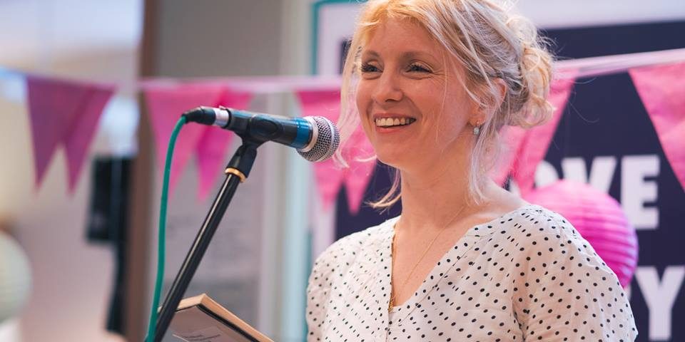 a blonde woman speaks into a microphone, smiling